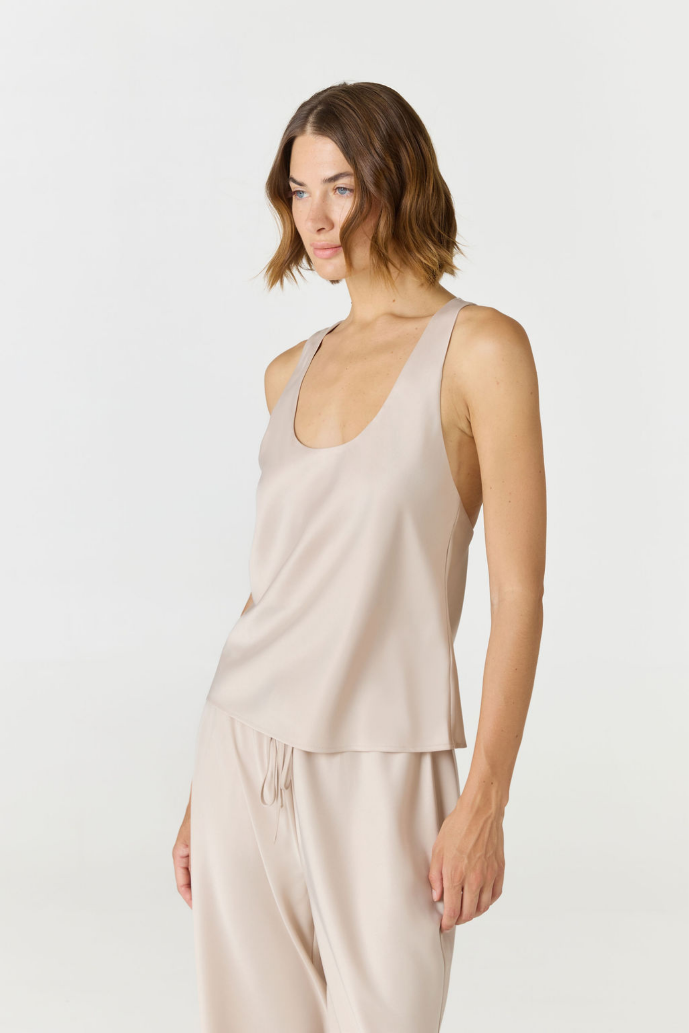 A woman modeling a sleeveless scoop-neck top and matching drawstring pants in a light beige or blush-taupe color, photographed against a plain light gray studio background.
