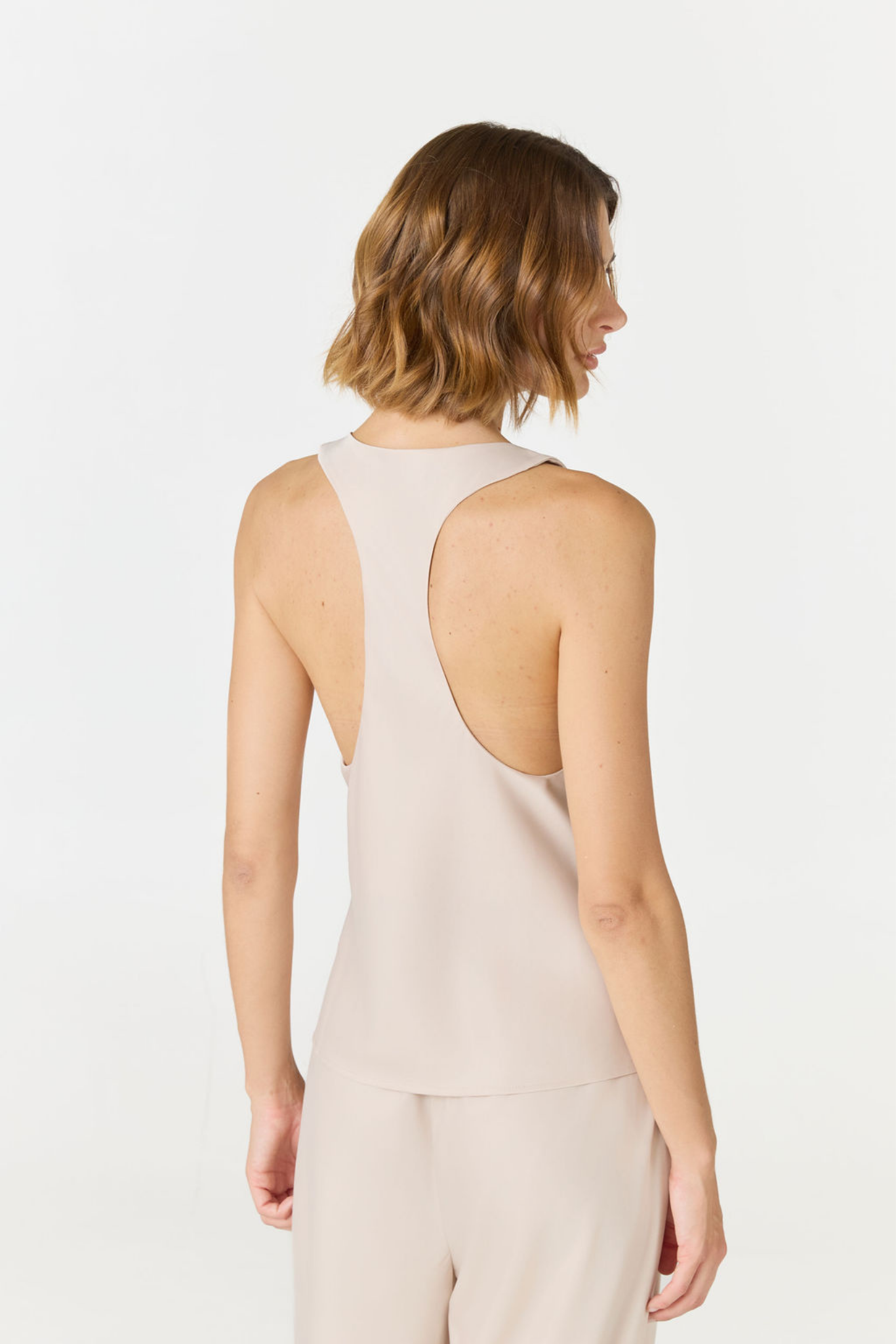 A woman shown from the back wearing a sleeveless light beige top and matching pants against a plain white studio background.