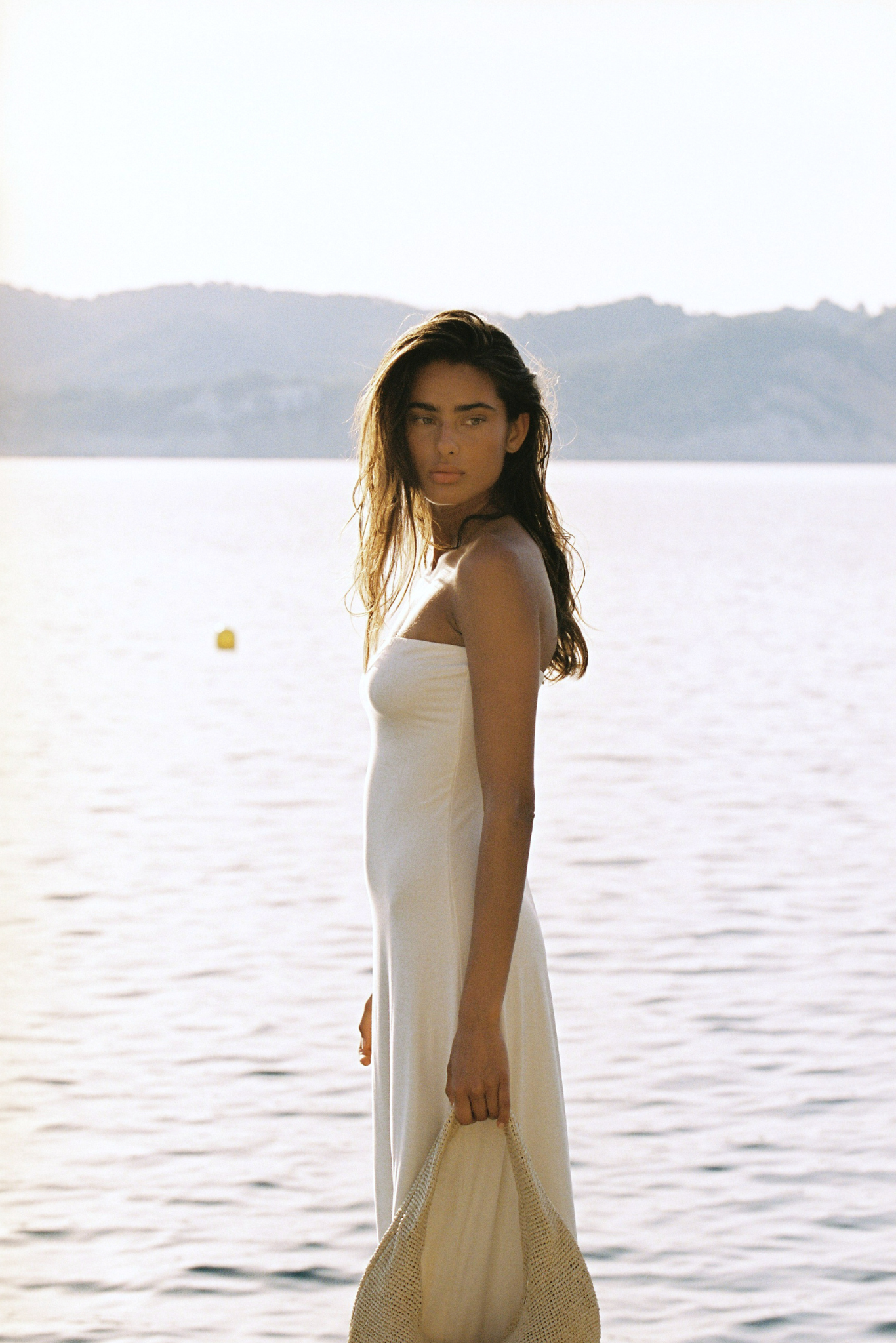 A woman stands by a calm body of water wearing a sleeveless white dress and holding a straw hat, with hazy hills in the background.