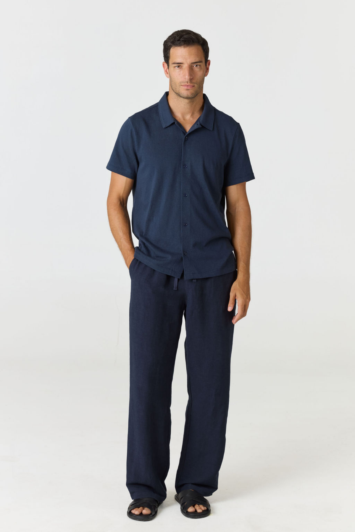 A man modeling a matching navy blue short-sleeve button-up shirt and wide-leg trousers set, standing against a plain light gray studio background.