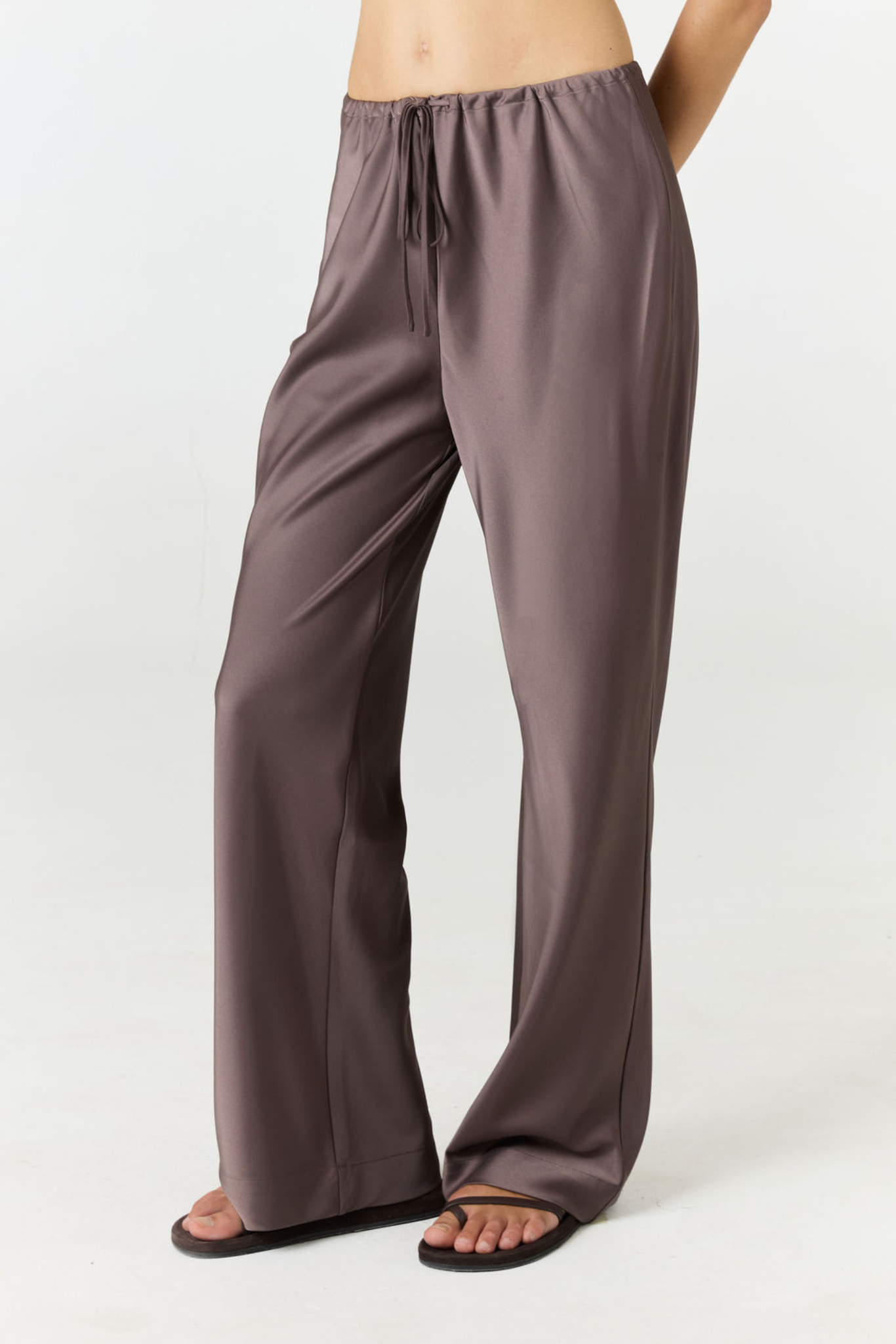 A person wearing wide-leg drawstring pants in a muted taupe-brown color, paired with black flip-flop sandals, shown from the waist down against a plain white studio background.
