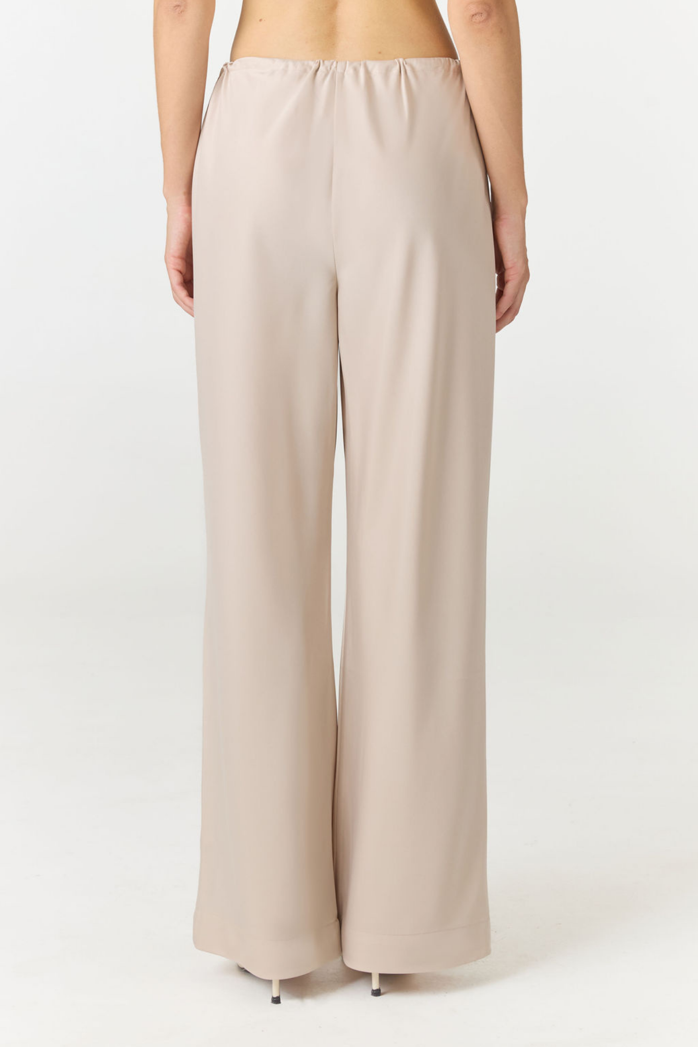 A model shown from the back wearing high-waisted, wide-leg trousers in a light beige or taupe color, paired with black heels, against a plain white studio background.