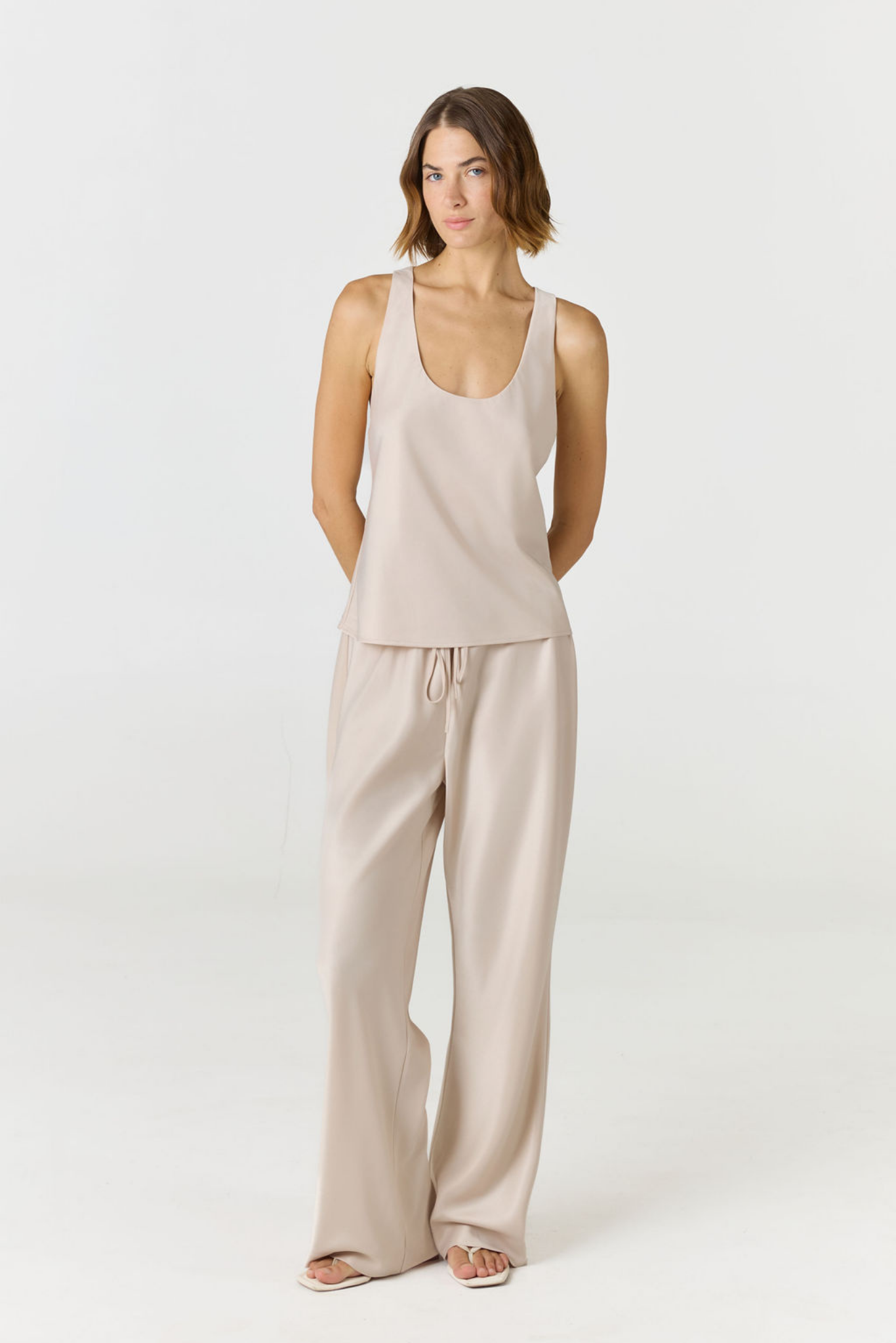 A woman modeling a light beige two-piece lounge set consisting of a sleeveless scoop-neck tank top and matching wide-leg drawstring pants, photographed against a seamless white studio background.