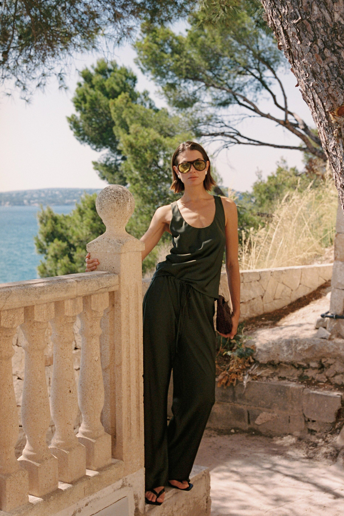 A woman wearing a dark olive green sleeveless jumpsuit and sunglasses stands on a stone terrace with a sea and trees in the background.