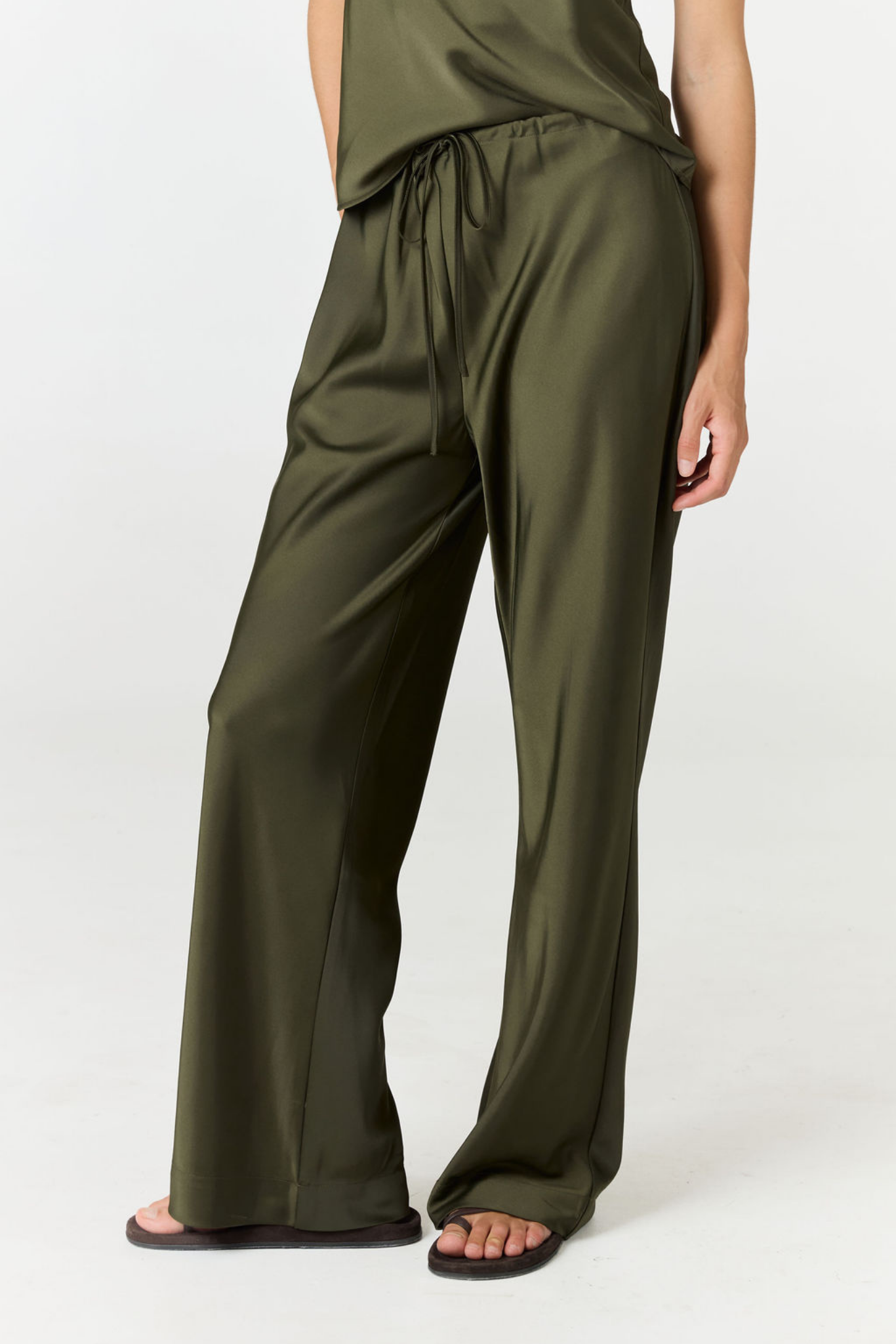 A person wearing olive green wide-leg satin pants with a drawstring waist, paired with dark sandals, shown against a plain light gray studio background.