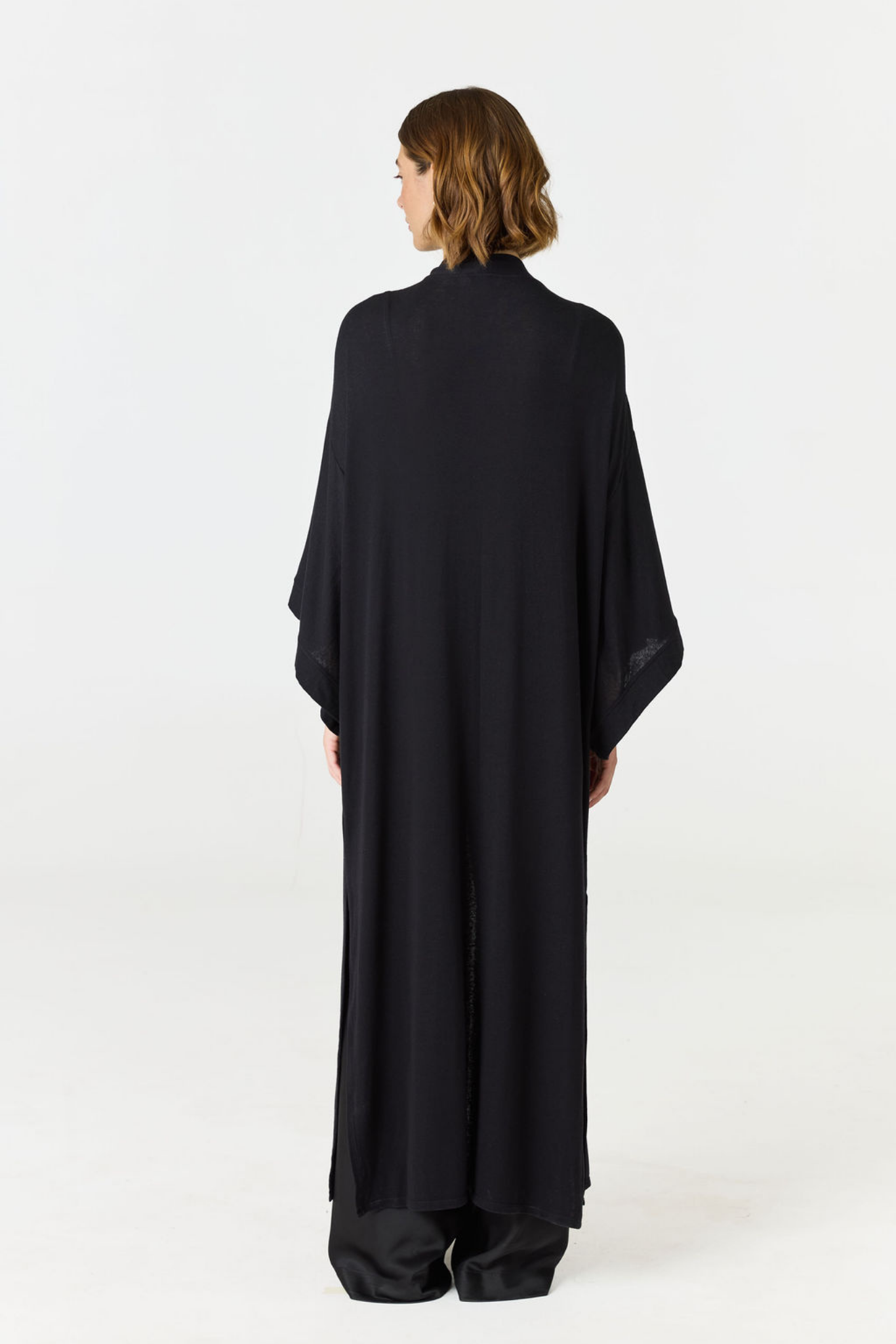 A model viewed from the back wearing a long, loose black abaya or caftan with wide draped sleeves, photographed against a seamless white studio background.