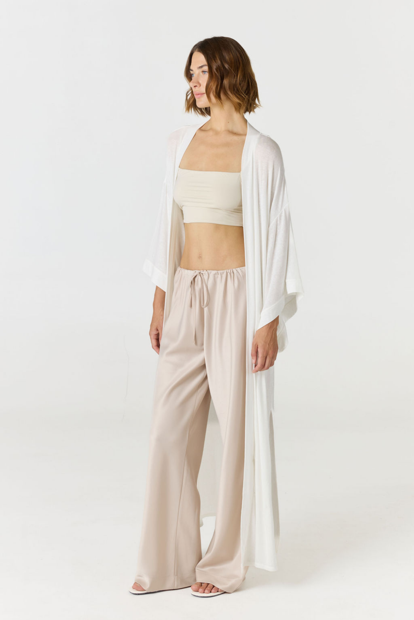 A woman models a three-piece neutral outfit consisting of a cream strapless tube top, light beige wide-leg drawstring pants, and a flowing white open-front cardigan, set against a plain white studio background.