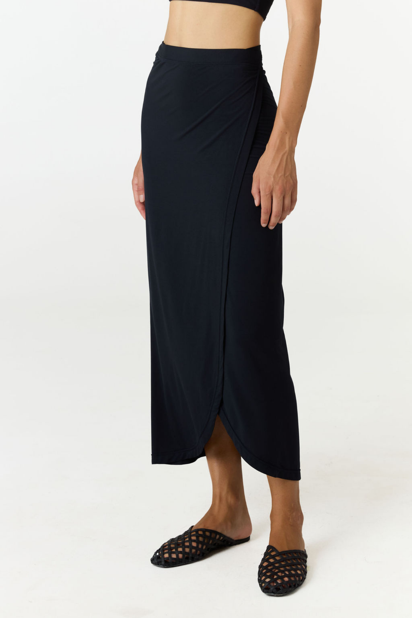 A model wearing a dark navy wrap midi skirt with a side slit, paired with black woven mule sandals, against a plain white studio background.