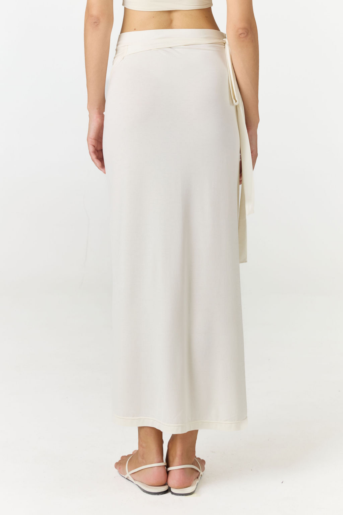 A model shown from the back wearing a cream-colored wrap maxi skirt with a side tie detail, paired with white strappy flat sandals against a plain white studio background.