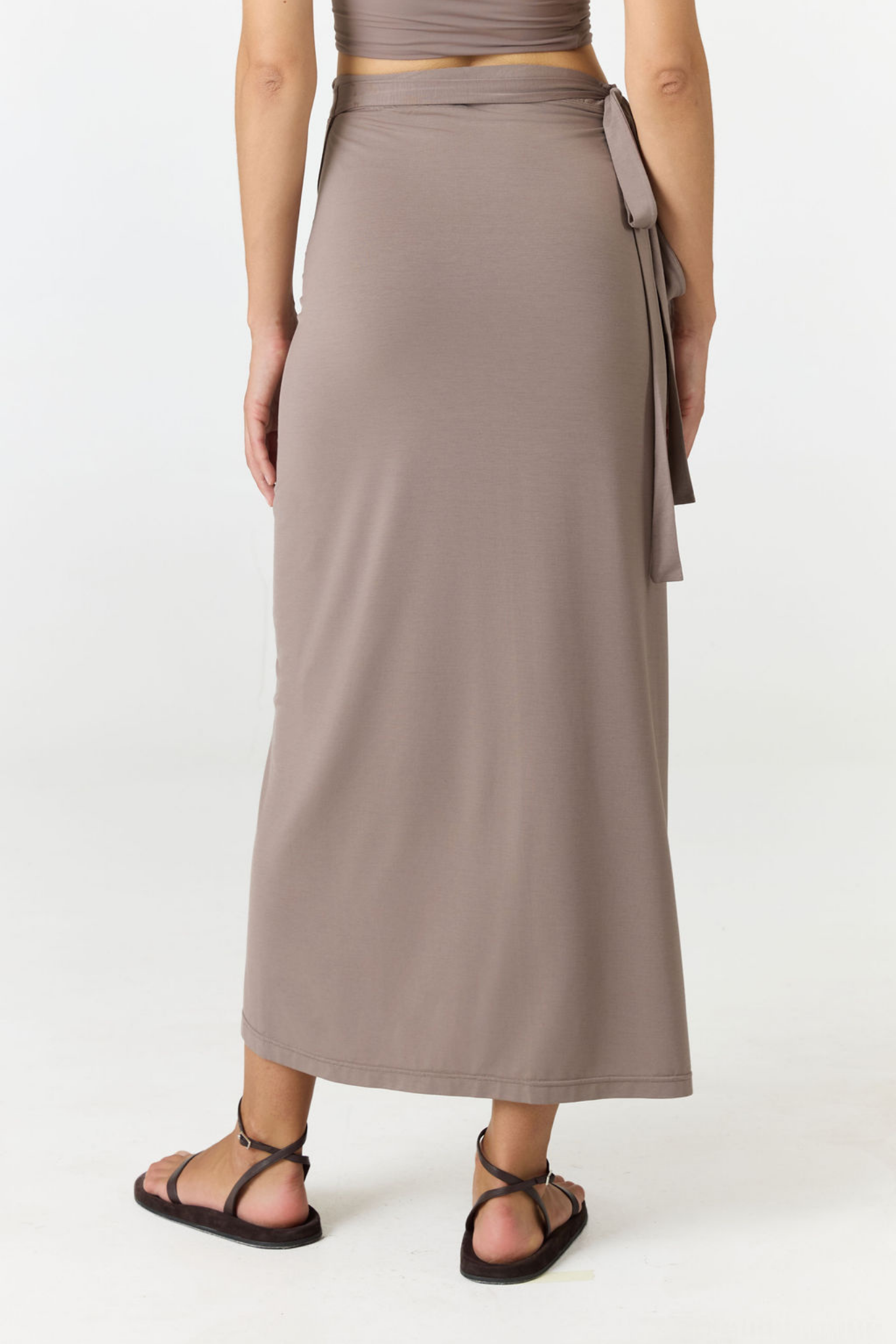 A woman wearing a taupe wrap maxi skirt with a side tie, paired with black strappy sandals, shown from the waist down against a plain white studio background.