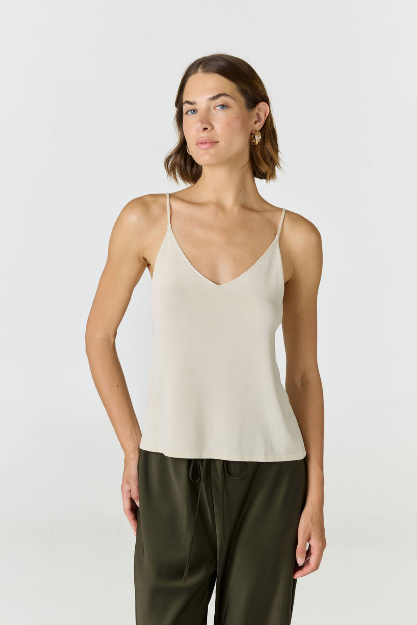 A woman modeling a light beige spaghetti-strap camisole top paired with dark olive green drawstring pants, shot against a seamless light gray studio background.
